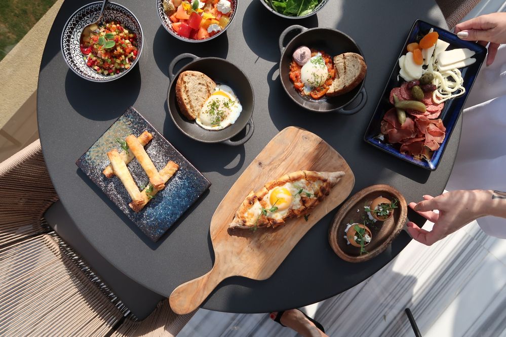 Caption: A photo of a round, black table filled with different dishes in bowls, plates, and a cutting board from Ruya Dubai, a restaurant in Dubai.