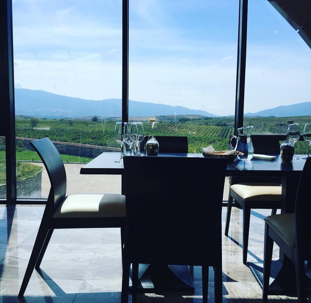 Billedtekst: Fem stole om et bord med vinglas og en kande vand, ved et stort vindue med udsigt over vinmarker og bjerge på Orbelus Vingård, Bulgarien. (Local Guide @MortenSI)