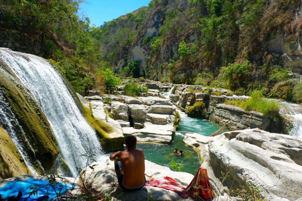 Caption: A photo of the back view of a person sitting at Tanggedu Waterfall with two people in the waters below. There are rocks and greenery all around. (Local Guide @MingguTerus).