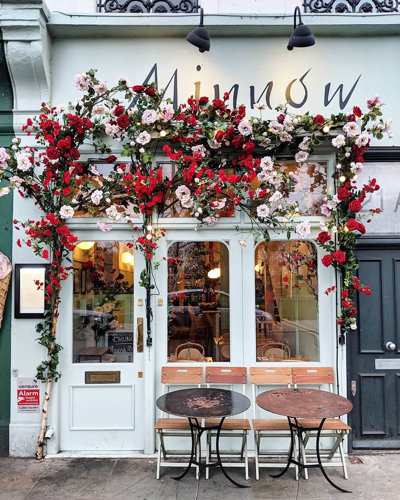 Caption: A photo of the exterior of Minnow, a restaurant in London, covered in pink and red flowers with two tables and four chairs outside of it. (Local Guide Bella Foxwell)