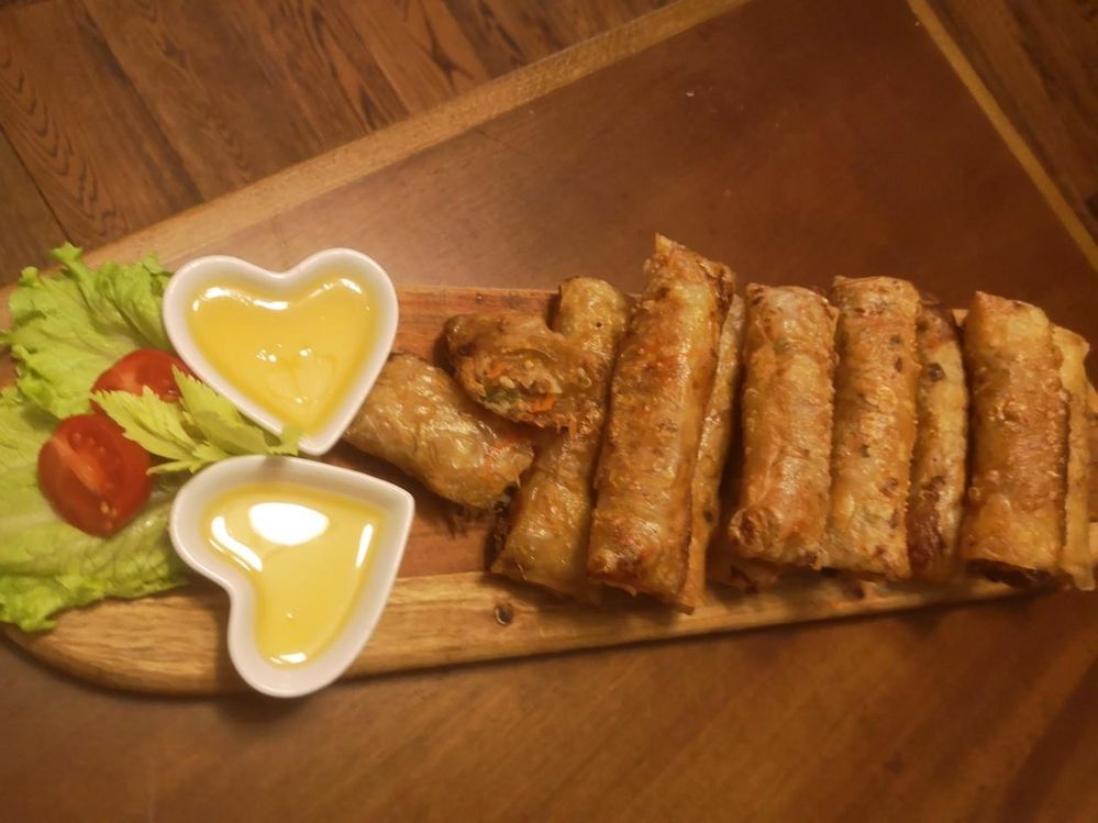 Caption: A photo of fried spring rolls with two small, heart-shaped bowls of yellow sauce for dipping and a cabbage leaf and a tomato for decoration on an oblong plate. (Local Guide @Aruni)
