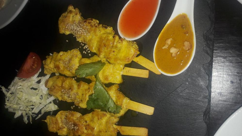 Caption: A photo of four chicken satay sticks, two deep spoons of peanut sauce, and sweet and sour sauce, and cut cabbage for decoration, served on a black plate. (Local Guide @Aruni)