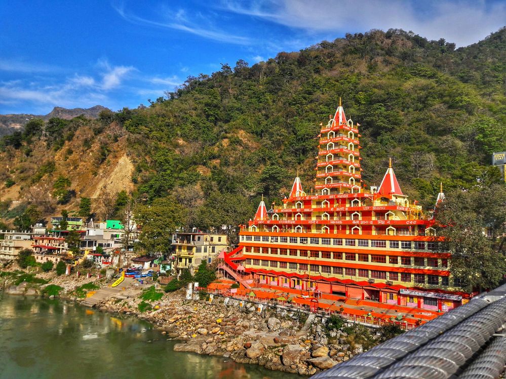 Caption: A photo of a red temple, houses, and mountains along the Ganges River in India. (Local Guide @IshantHP_ig)