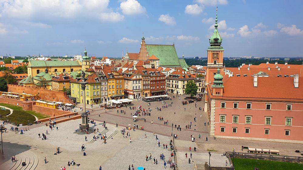 Caption: A photo of the Plac Zamkowy Square in the old town of Warsaw. It shows many buildings and people walking around the square. (Local Guide @christophesubilia)