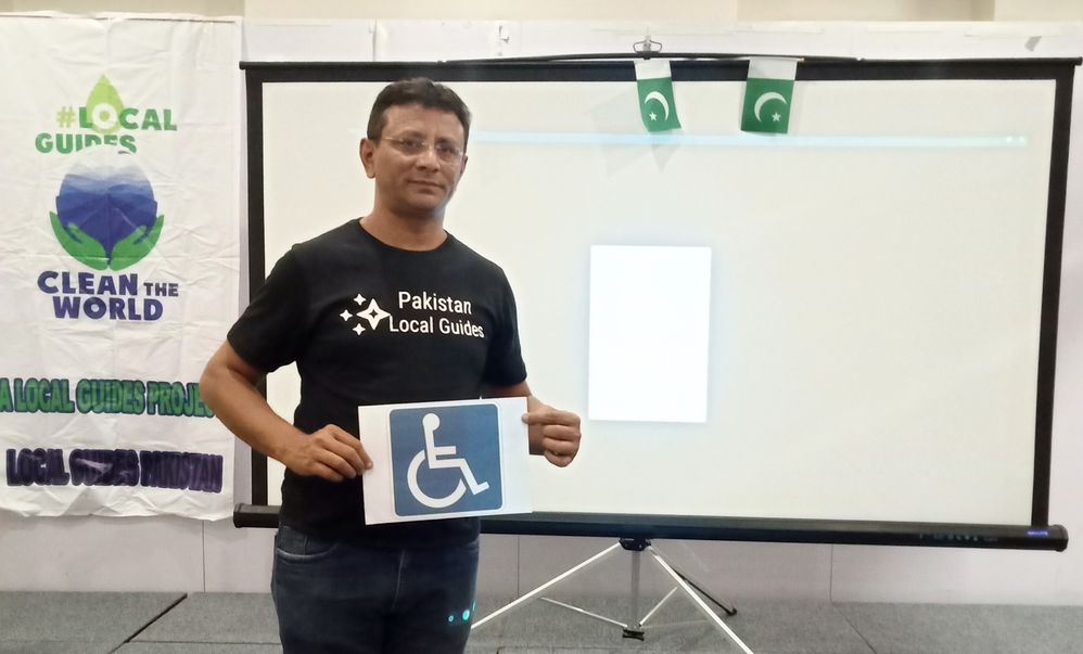 Caption: A photo of Local Guide Kashif Abdul Pazzaq holding a sign with the accessibility icon on it and wearing a shirt that says “Pakistan Local Guides.” (Courtesy of Local Guide @Kashifmisidia)