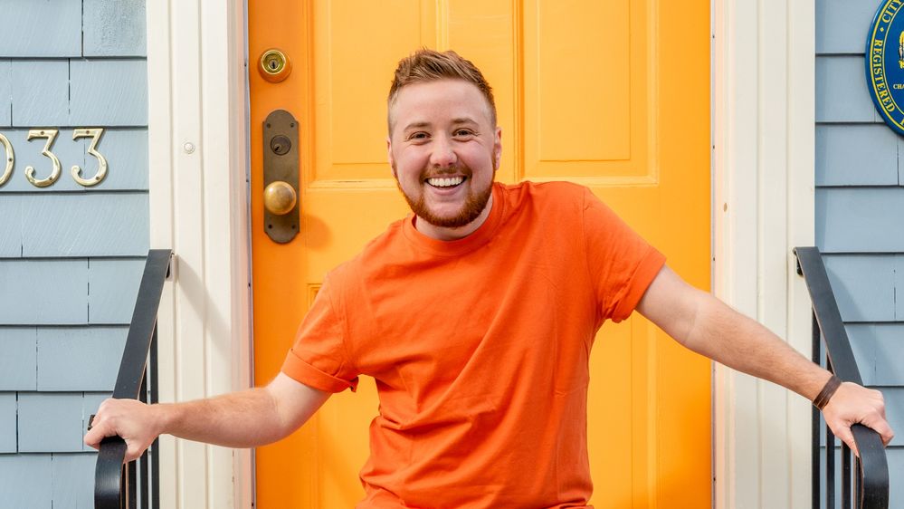 Caption: A portrait photo of Local Guide Samson L. wearing an orange shirt and standing in front of an orange door. (Aaron Mckenzie Frazier)