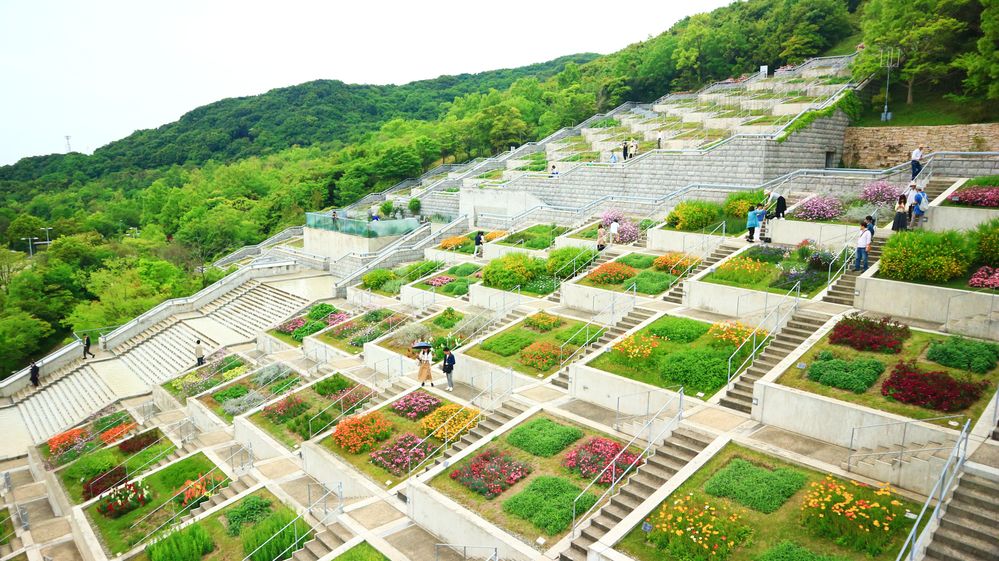 Caption: A photo of multiple square flower beds arranged in grids and spread over several levels on a hill, with staircases between them at Awaji Yumebutai in Awaji, Japan. (Local Guidе 美濃英樹)