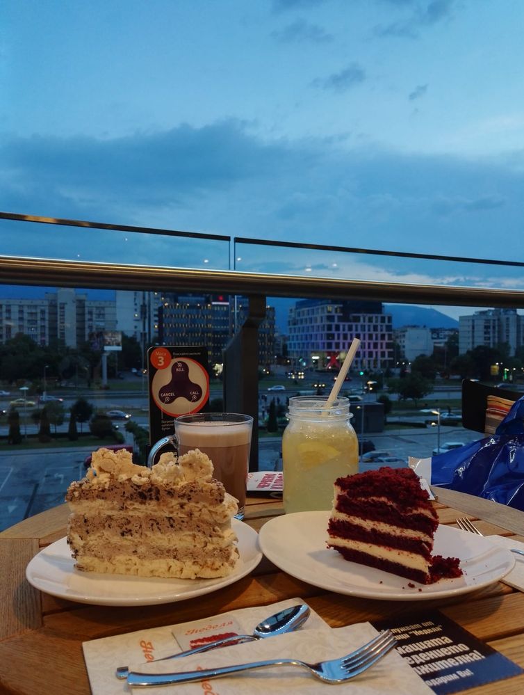 Caption: A photo of two cakes, a cake full of meringues on the left and a red velvet cake on the right with two drinks on the background and a nice view after sunset towards the mountain. (Local Guide @TsekoV)