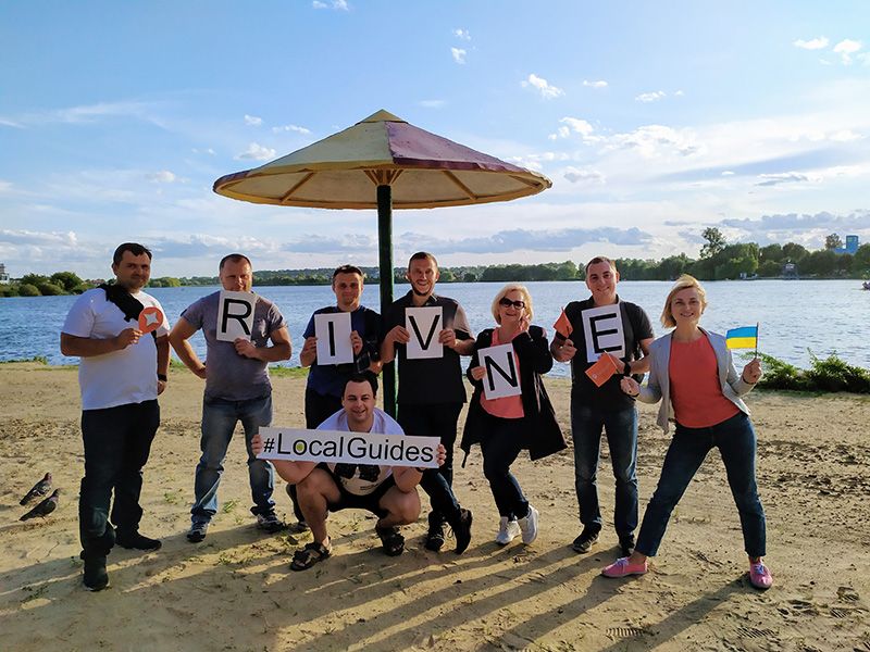 Caption: A photo of eight Ukrainian Local Guides standing in front of a lake and holding the Ukrainian flag, a poster with the #LocalGuides, and paper letters that spell the name “Rivne”. (Local Guide @NatalkaR)