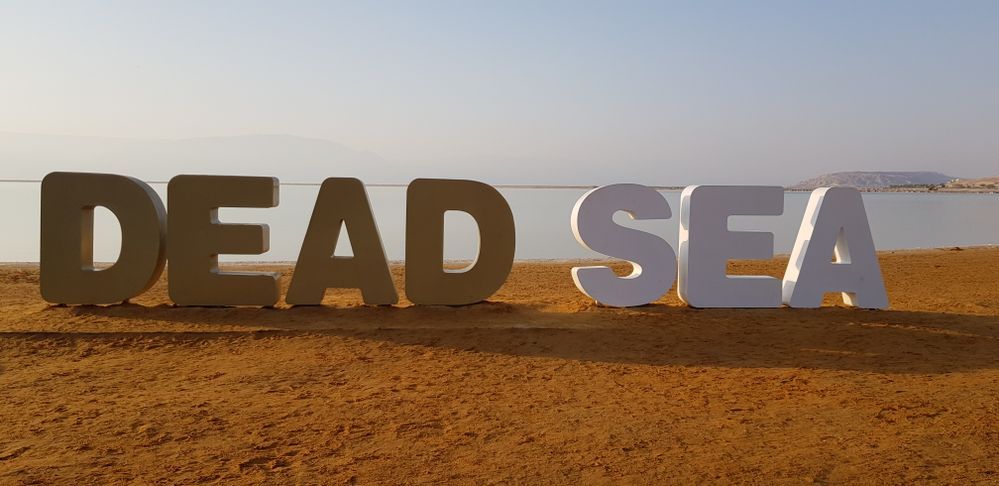 Caption: A photo of three dimensional letters that spell “Dead Sea”, placed on the brown sand beach in front of the Dead Sea in Israel. (Local Guide @Avsha)