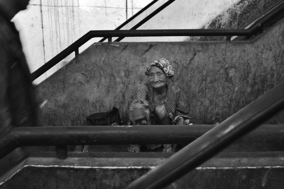 Caption: A black and white photo of an old woman sitting on the street next to a staircase. (Local Guide @sansa)