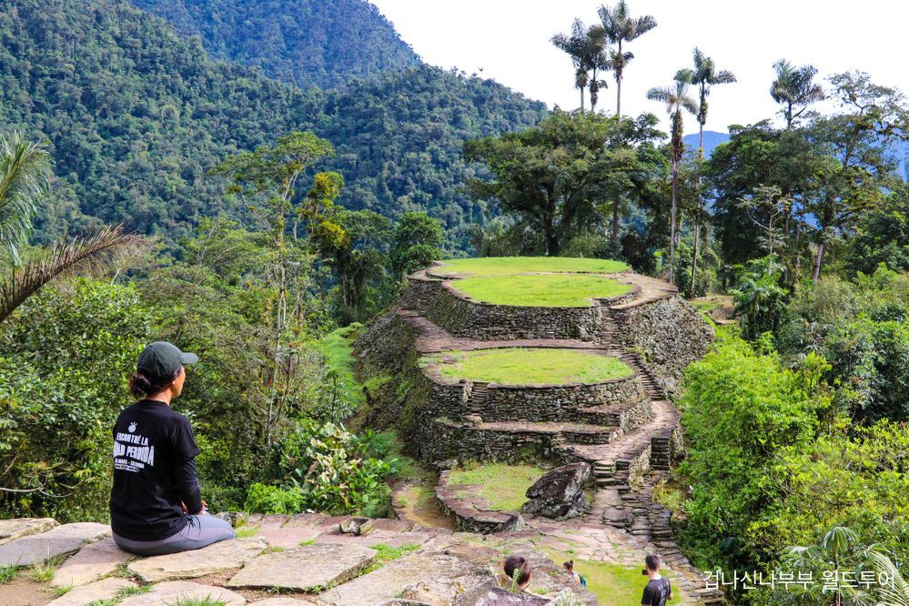 Caption: A photo of a woman and a dog looking at the archaeological site of the Lost City in Colombia, surrounded by lush green mountains. (Local Guide @겁나신나부부)