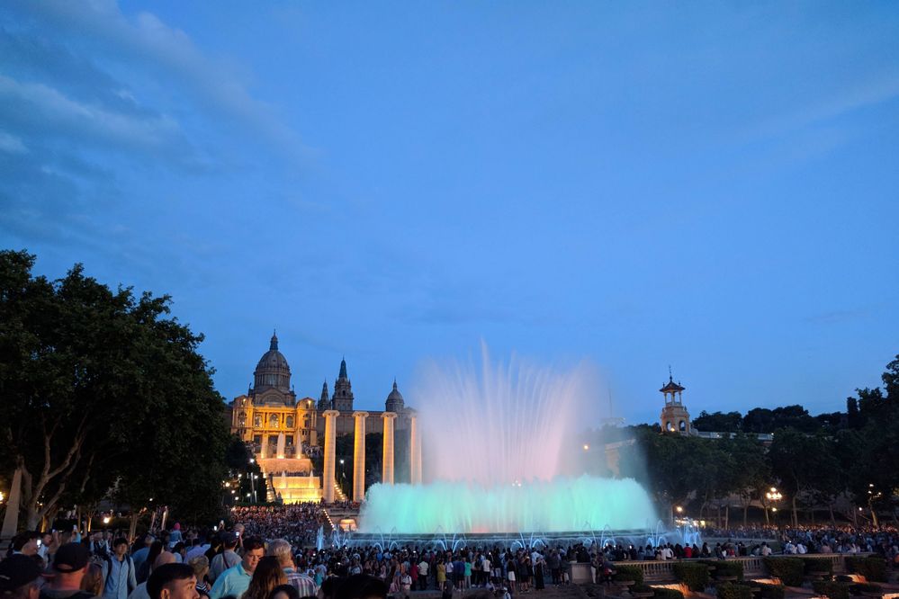 Caption: A photo of the Magic Fountain of Montjuic in Barcelona, Spain, lit up in a turquoise colour and surrounded by a lot of people, with the National Museum of Art in the background. (Local Guide @MoniDi)