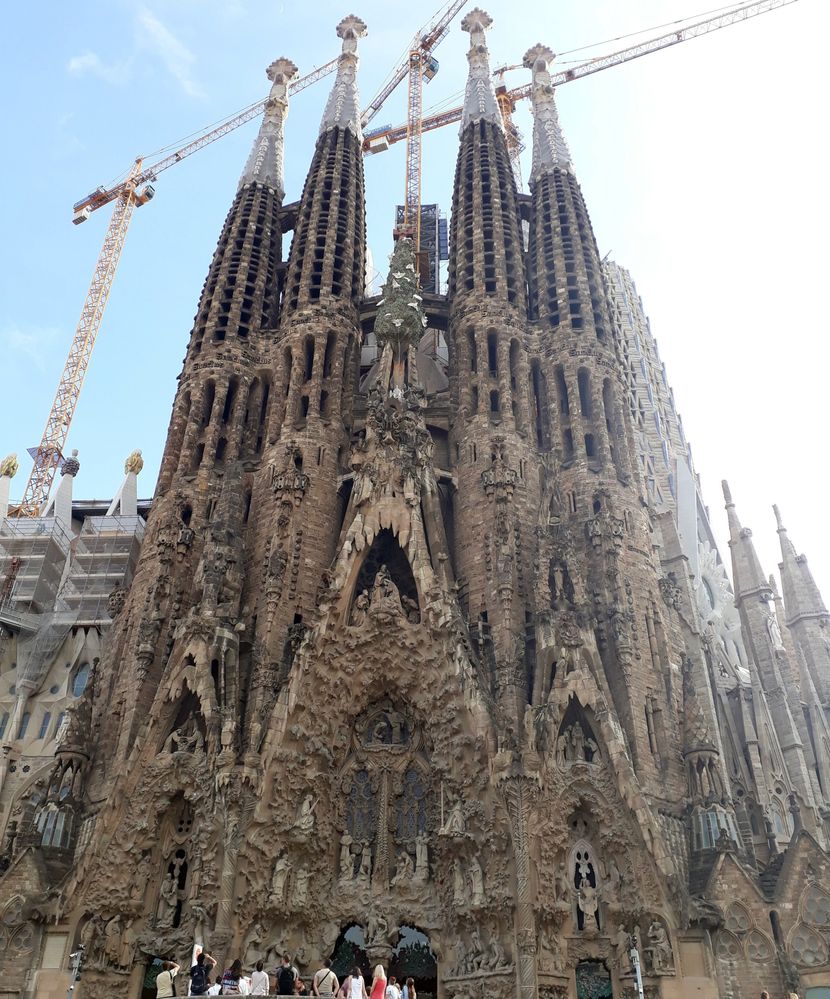 Caption: A photo of the facade of the Sagrada Familia Cathedral with its several steeples and cranes towering over them in the background. (Local Guide @MoniDi)
