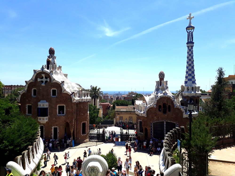 Caption: A photo of the main gate of Park Guell, with Casa del Guarda on the left and the official souvenir shop on the right. Part of Barcelona and the Balearic Sea can be seen in the distance. (Local Guide @MoniDi)