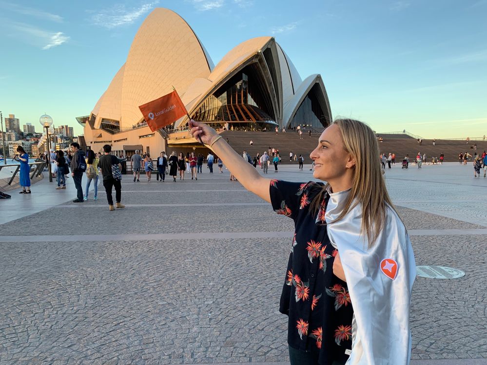 Caption: A photo of Penny Christie wearing a homemade Local Guides cape and holding a Local Guides flag in front of the Sydney Opera House in Sydney, Australia. (Local Guide @PennyChristie)