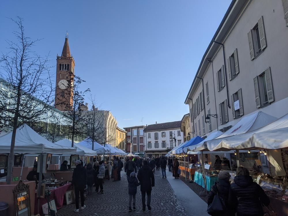Caption: A photo of a street lined with stalls underneath white sunshades in Treviglio, Italy. There are people walking around and looking at the antiques on sale. (Local Guide @RiccyB)