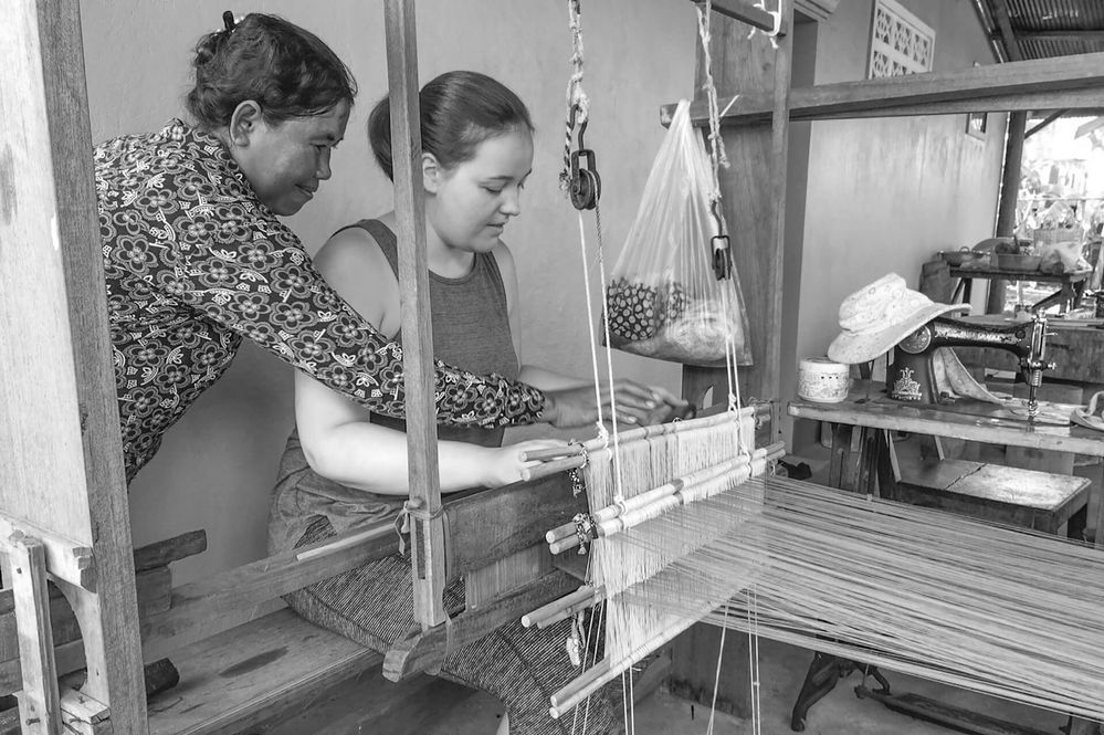 Caption: A black and white photo of Local Guide @mhcouette learning how to weave using a handloom and a Cambodian woman giving her advice. (Local Guide @mhcouette)