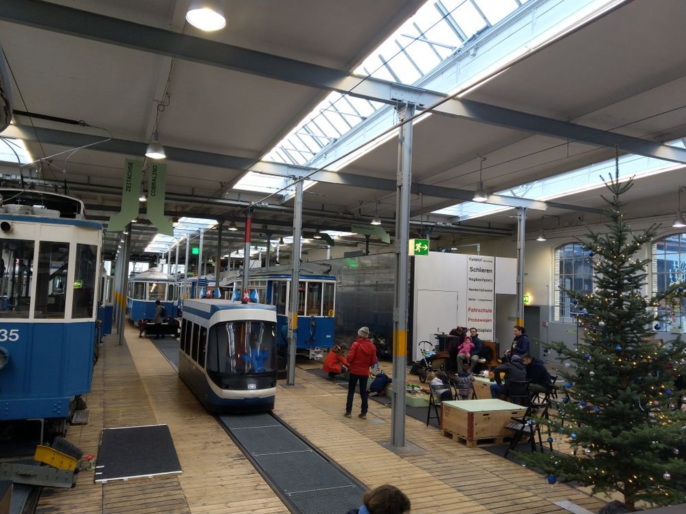 Caption: A photo inside the Tram Museum in Zurich, Switzerland, showing several trams of different sizes, a Christmas tree, and parents with children relaxing on chairs. (Local Guide @KseniaM)