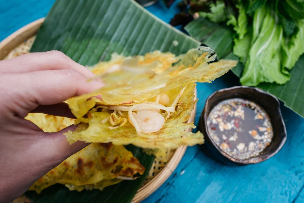 Caption: A photo of a hand holding a crispy rice flour crepe filled with shrimp and sprouts. There’s a small bowl of fish sauce on the side. (Local Guide Piotr Górecki)