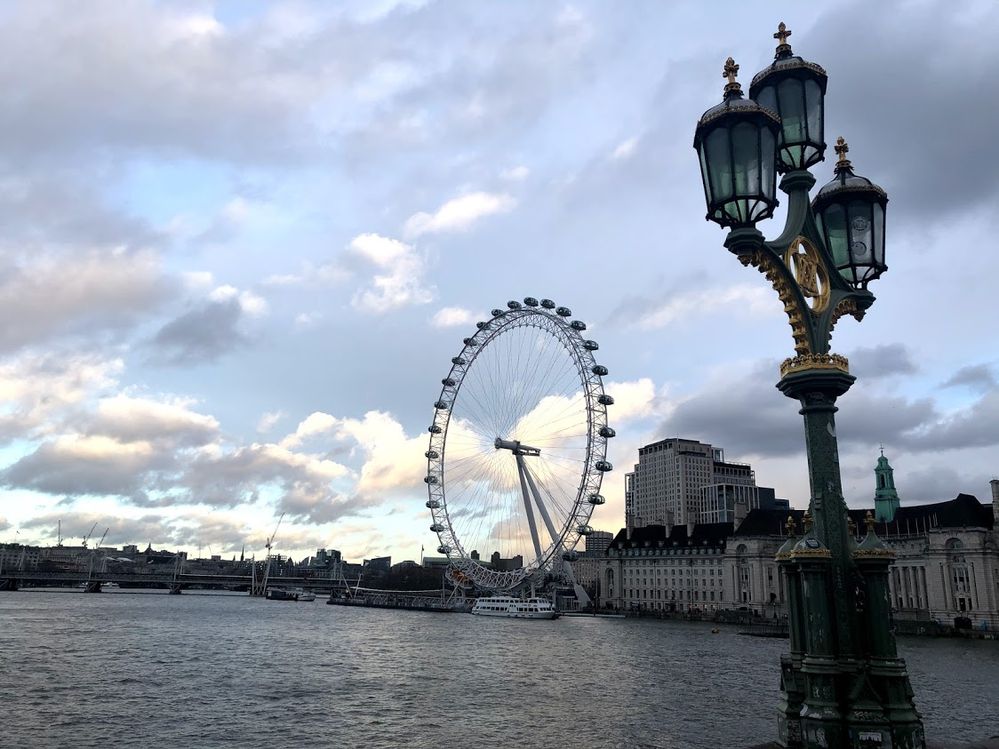 Legenda: Um foto da London Eye tirada da ponte Westminster mostrando a roda-gigante, alguns prédios ao fundo, o Rio Tâmisa com algumas embarcações e um lustre muito bem decorado. (Local Guide @ItzPirk)