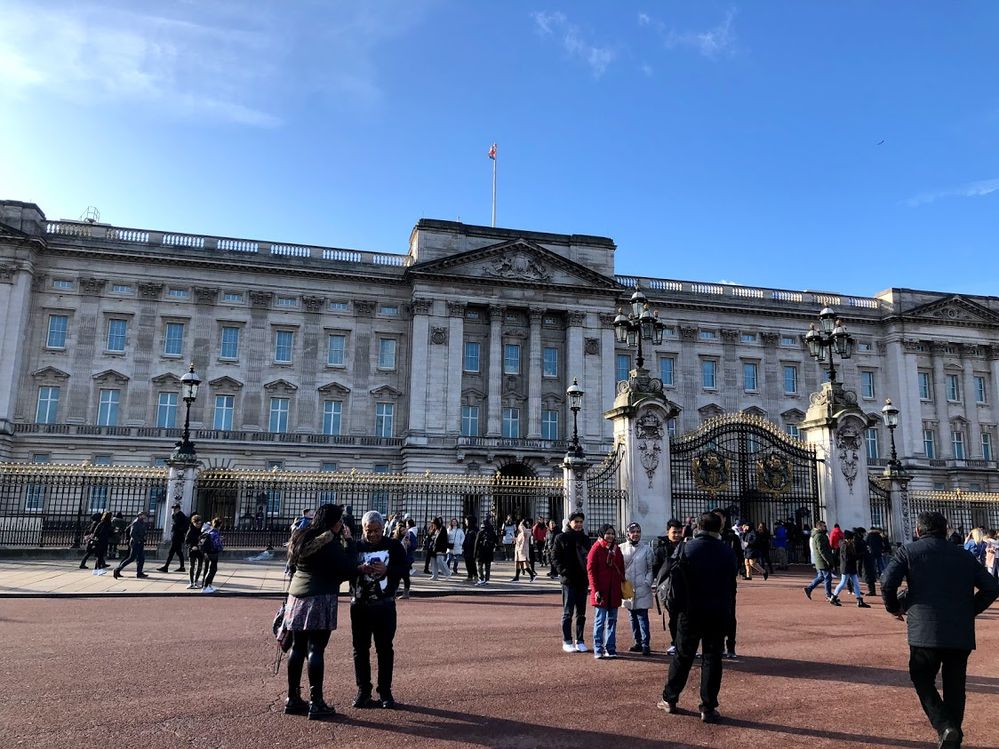 Legenda: Uma foto da frente do Palácio de Buckingham mostrando o prédio bem detalhado com seu portão com detalhes em dourado com alguns turistas a frente dele. (Local Guide @ItzPirk)