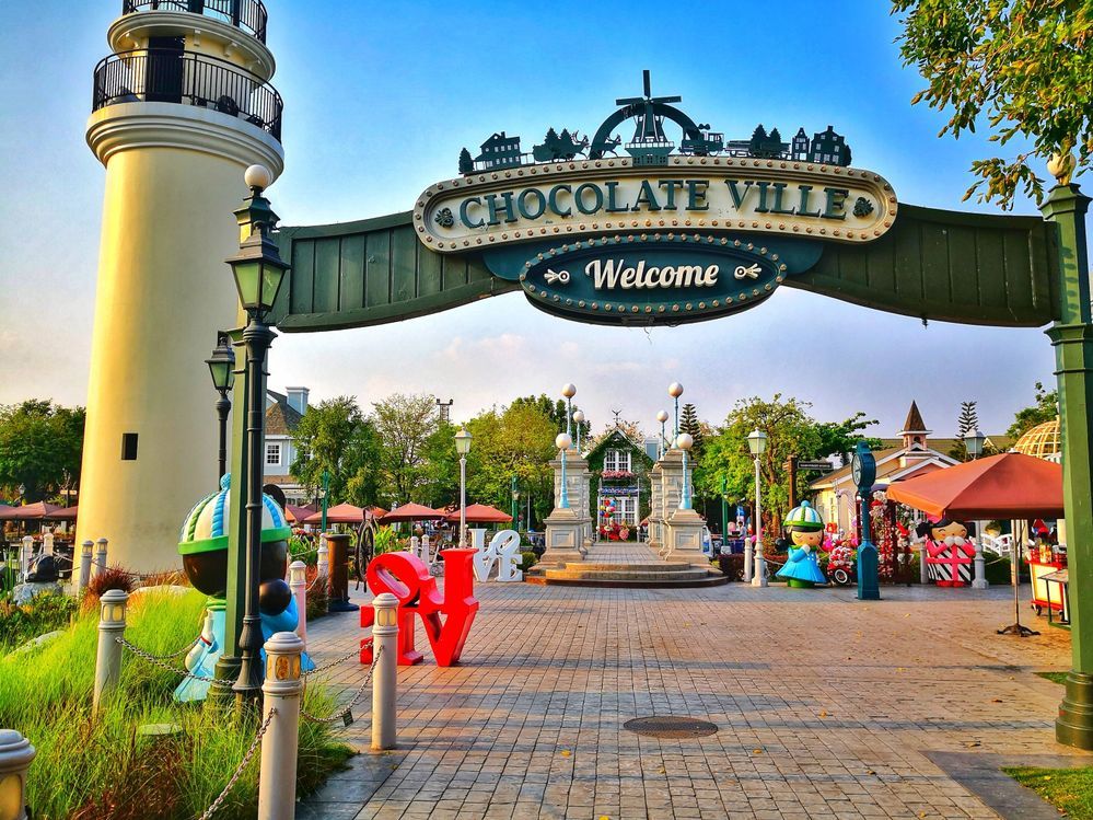 Caption: A photo of the entrance to Chocolate Ville in Thailand, showing a green metal arch with a “welcome” sign, and a beautiful ornamental tower and model houses in the background. (Local Guide @GoldenPapa)