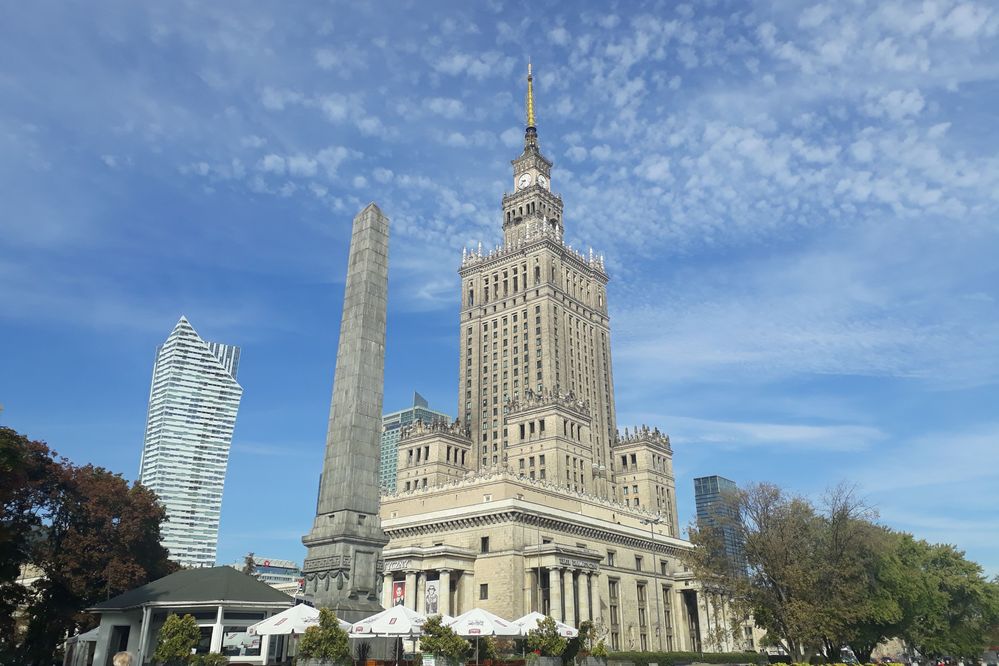 Caption: A photo of the imposing Palace of Culture and Science in Warsaw, with a monument in front of it, and a tall building in the background. (Local Guide @MoniDi)