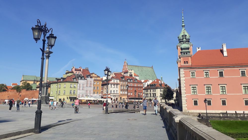 Caption: A photo of a square and several colorful buildings in the Old Town of Warsaw, with the reddish facade of the Royal Castle on the right. (Local Guide @MoniDi)