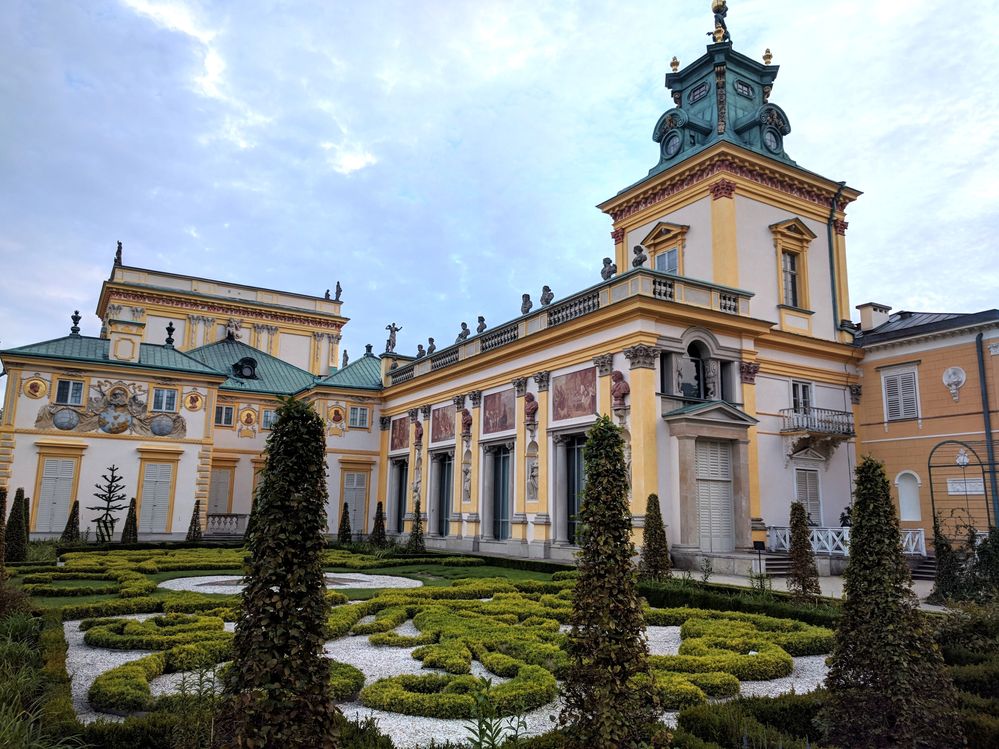 Caption: A photo of a lush green Baroque garden and a part of the Wilanow Palace with its copper roof. (Local Guide @MoniDi)