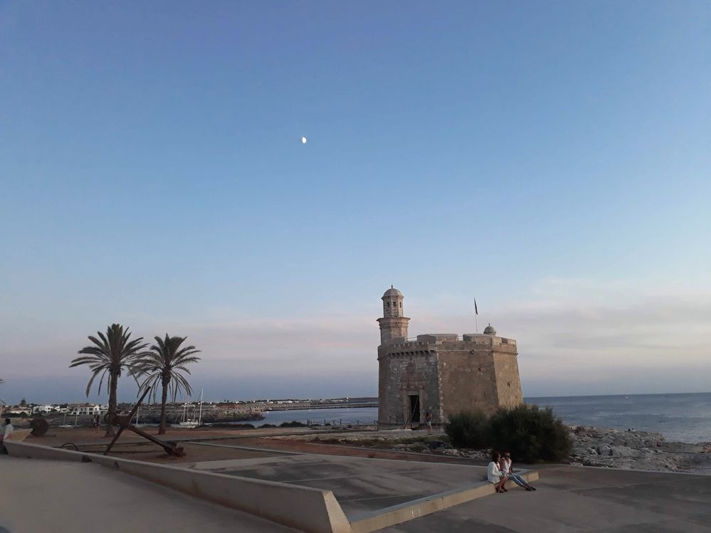 Caption: An evening photo of the Castell de Sant Nicolau stone tower, two palm trees, and the harbor in Ciutadella de Menorca. (Local Guide @MoniDi)